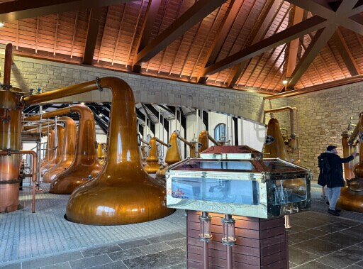 Glenfiddich distillery binnen: je ziet grote koperen distilleerketels in een oud gebouw met houten constructiedak. Op de voorgrond staat een kastje met gouden deurtje. 
